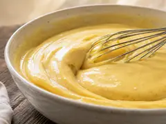 Crema pastelera amarilla y brillante en un bol de ceramica, espesa y con puntos de vainilla, con un batidor de mango de madera apoyado adentro, sobre mesa de madera
