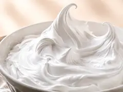 Merengue italiano blanco y brillante en un bol de ceramica, con picos altos que se curvan en la punta y el batidor apoyado al costado, sobre mesa de madera