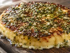 Provoleta a la parrilla dorada sobre tabla de madera, con provenzal de ajo y perejil en aceite por encima