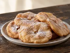 Tortas fritas caseras apiladas en un plato de cerámica clara sobre mesa de madera oscura, espolvoreadas con azúcar impalpable