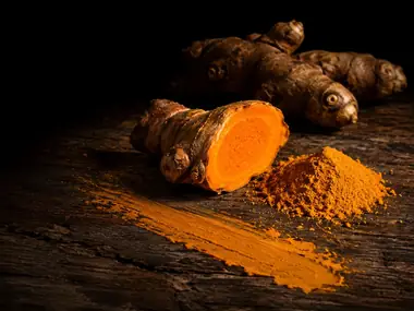 Rizomas de cúrcuma sobre madera, uno cortado mostrando el naranja intenso, junto a un montoncito de cúrcuma molida
